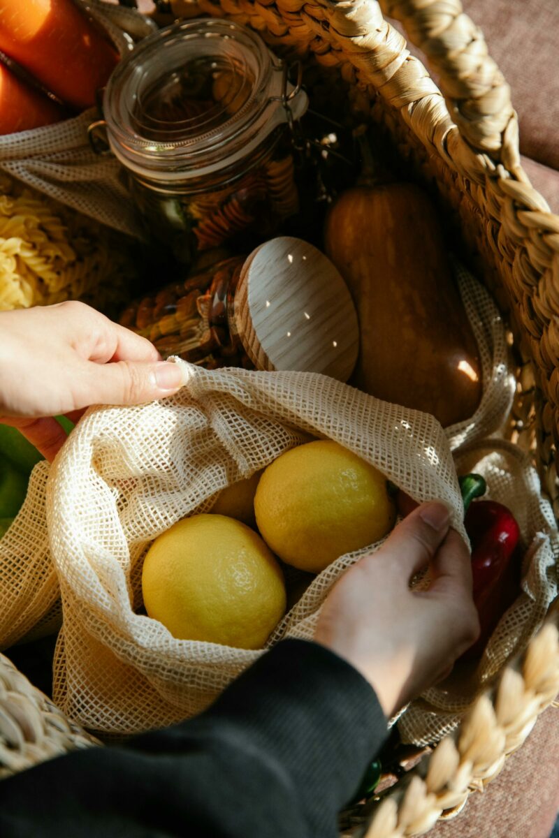 From above of crop anonymous person with bag full of fresh lemons placed in wicker basket with jars of assorted food